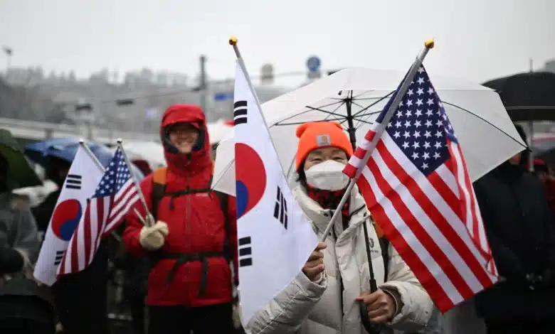 Un groupe de personnes se tient dehors sous la neige, tenant des drapeaux sud-coréen et américain. Certains portent des vêtements chauds et des masques, et quelques-uns tiennent des parapluies pour se protéger de la neige. Latmosphère semble festive et animée.