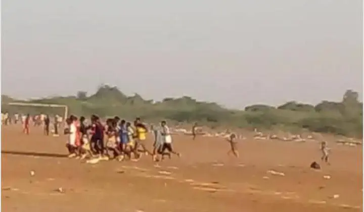 Un groupe de personnes joue au football sur un vaste terrain poussiéreux avec un poteau de but en arrière-plan. Une végétation clairsemée et des individus dispersés peuvent être vus au loin sous un ciel clair.