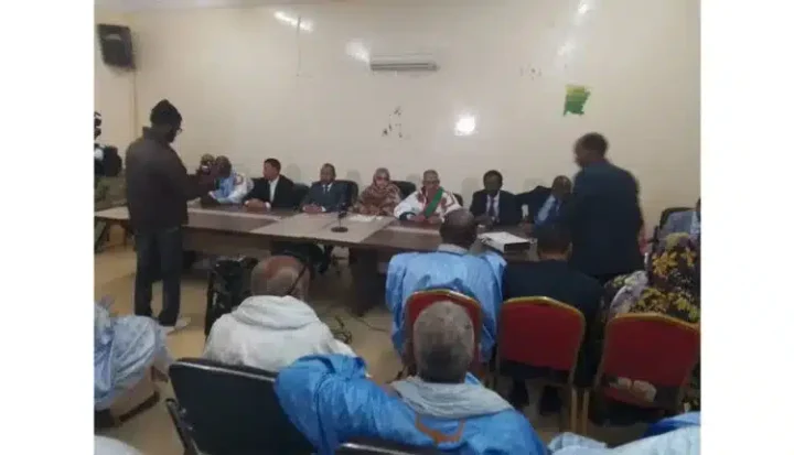 Un groupe de personnes est assis à une longue table dans une salle de conférence, face à un public. Un homme vêtu de vêtements sombres se tient près dune caméra à gauche, tandis quun individu en tenue traditionnelle marche à droite. Certains participants portent des vêtements bleu clair.