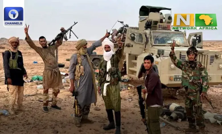 Un groupe dhommes armés se tient dans une zone désertique, posant devant un véhicule militaire. Certains tiennent des armes et font des signes de victoire avec leurs mains, rappelant les rapports sur les « civils tués au Mali ». Des objets éparpillés jonchent le sol tandis que les logos de Channels TV et de Network Africa apparaissent dans les coins.