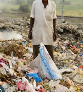 Une personne se tient debout au milieu dun grand tas dordures, tenant un sac et un outil. La zone est remplie de différents types de déchets, notamment du plastique et du papier. Larrière-plan montre un ciel brumeux et une route.