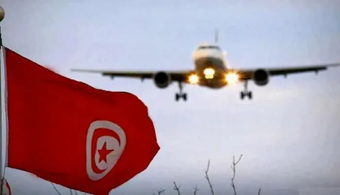 Un drapeau rouge avec un symbole en forme de croissant et détoile flotte au premier plan, tandis quun avion sapproche pour atterrir en arrière-plan, avec ses phares datterrissage allumés sur un ciel nuageux.