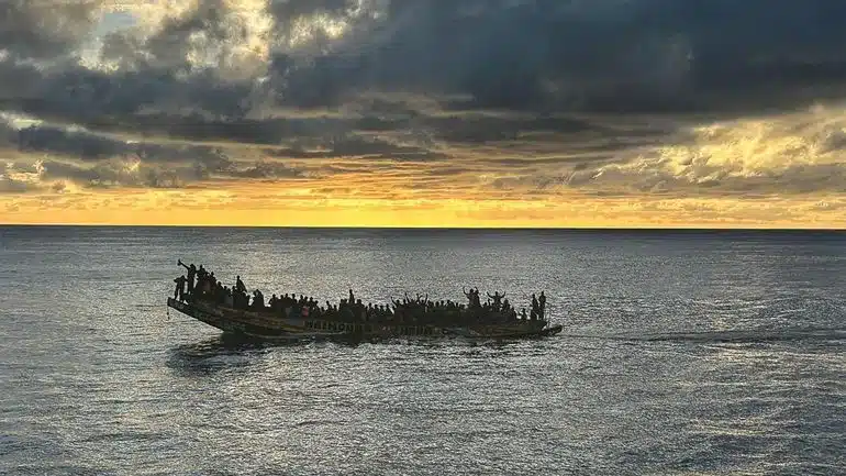 La silhouette dun bateau bondé de monde navigue sur un vaste océan sous un ciel dramatique. Le soleil se couche, projetant des teintes dorées sur leau et les nuages, créant un contraste saisissant entre la lumière et les ombres.