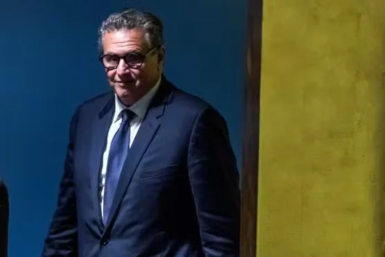 Un homme en costume sombre et lunettes se tient dans un espace faiblement éclairé, sur fond bleu et jaune. Il a les cheveux gris et regarde vers lavant avec un léger sourire.