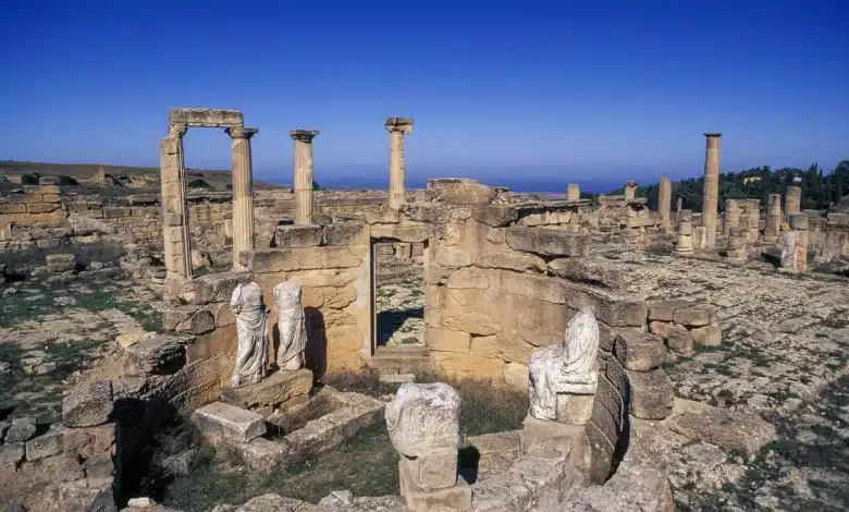 Ruines antiques sous un ciel bleu clair, avec des colonnes debout et brisées et des statues fragmentées. Le site est entouré dun terrain herbeux et des vestiges de murs en pierre sont visibles, créant une disposition circulaire.