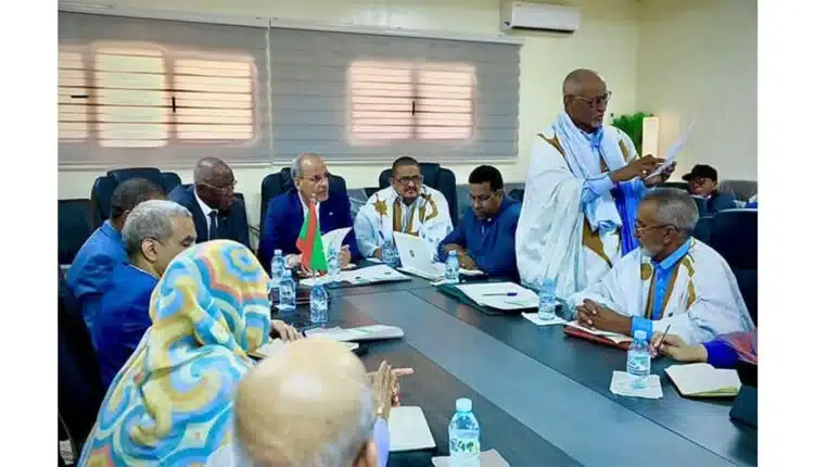 Au Brakna,un groupe de personnes en tenue traditionnelle et en costume sont assises autour dune table de conférence en pleine réunion. Certaines personnes ont des ordinateurs portables et des documents, et un drapeau est visible sur la table. La salle est bien éclairée avec de grandes fenêtres en arrière-plan.