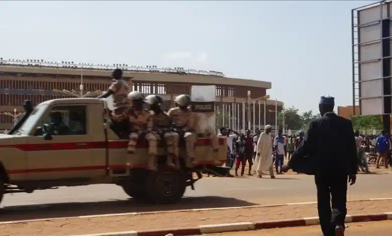 Un groupe de policiers en uniforme est assis à larrière dun camion de police en mouvement dans une rue. À proximité, des gens sont rassemblés et un homme en costume sombre marche dans la direction opposée. Un bâtiment moderne et des arbres sont visibles en arrière-plan sous un ciel clair.