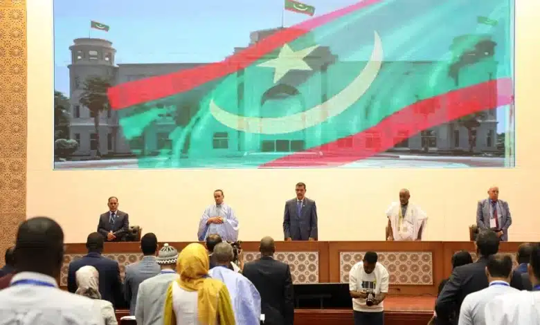 Participants debout lors de la cérémonie d’ouverture de la conférence nationale des médecins mauritaniens, devant un grand écran affichant le drapeau de la Mauritanie au Palais des Congrès de Nouakchott.