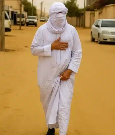 Mohamed Ould Cheikh El Ghazouani marchant incognito dans une rue sablonneuse de Nouakchott, vêtu d’un boubou blanc et d’un turban.
