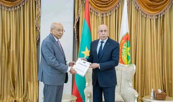 Deux hommes échangent un document lors d’une cérémonie officielle au palais présidentiel, devant les drapeaux mauritaniens.