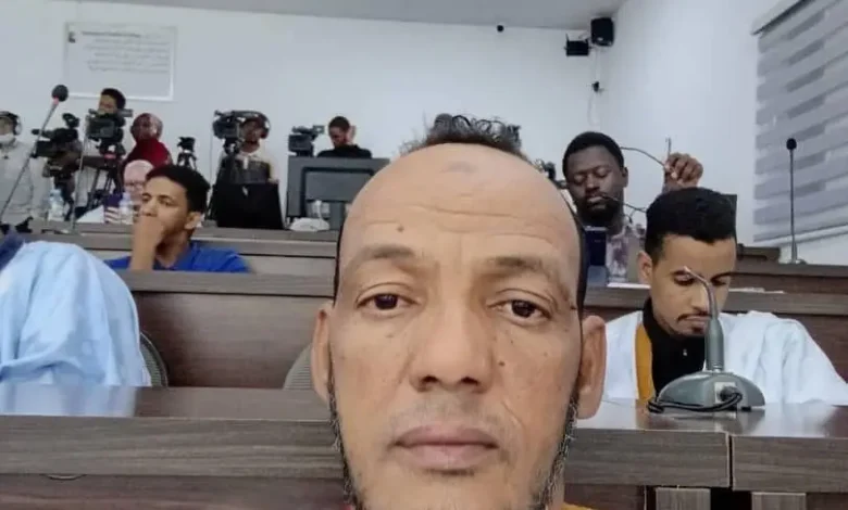 Un homme assis dans une salle de conférence lors d’une réunion ou séance officielle, entouré de journalistes et de caméras.