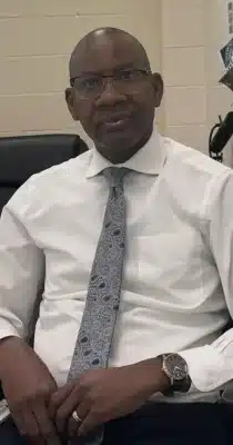 Homme assis dans un bureau, vêtu d’une chemise blanche et d’une cravate à motifs, portant des lunettes.